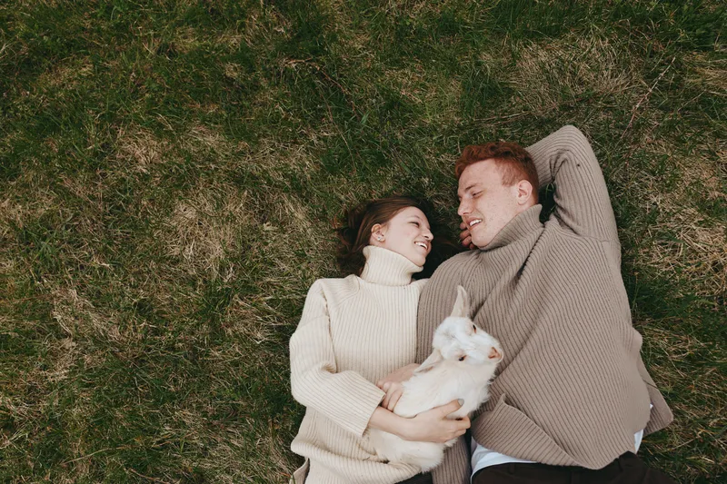 Couple lying on grass with a white goat kid, smiling and cuddling under soft daylight. Cozy sweater weather, pastoral lifestyle, intimate moment in nature, relaxed mood, gentle connection, tranquil