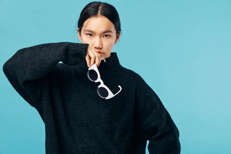 Young woman in black sweater posing with white sunglasses on blue background. Fashion portrait of confident female model with sleek hair and casual style for lifestyle or beauty concept.