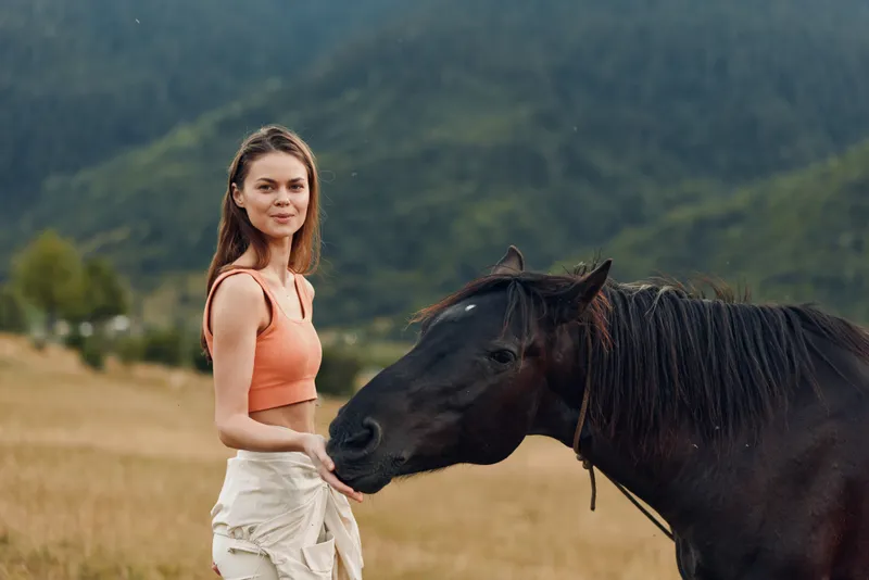 A confident woman stands beside a dark horse in an open field beneath distant mountains. She smiles softly, radiating calm, trust, and a harmonious bond with the animal.