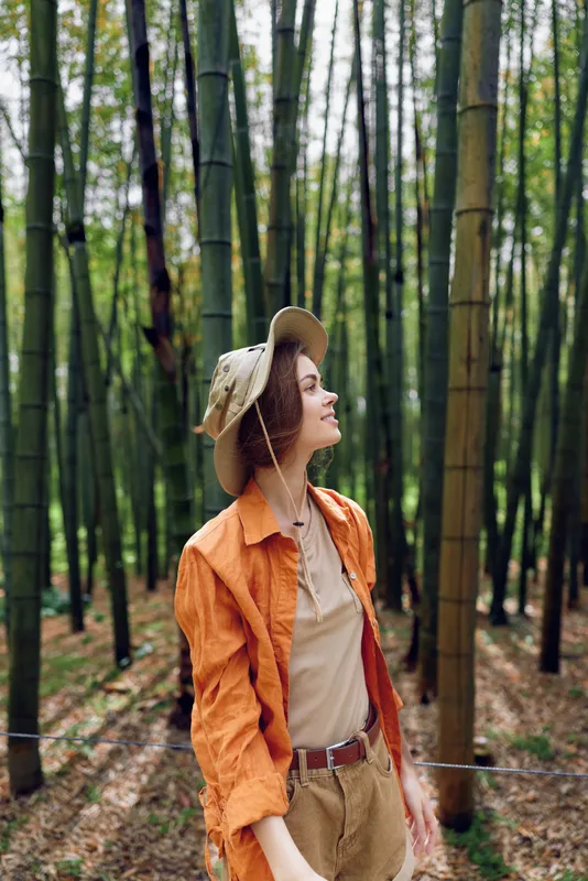 Woman in bamboo forest wearing straw hat and orange jacket, smile and relaxed nature portrait during travel, casual outfit and sunlit greenery among tall trees