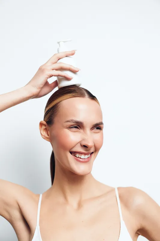 Smiling woman with clear skin holding white lotion bottle above her head, applying skincare product. Bright studio lighting emphasizes healthy skin and beauty care concept.