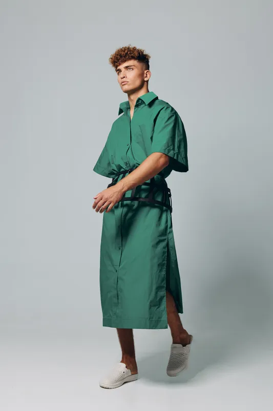Young man with curly hair wearing green oversized shirt dress and white sneakers standing against plain background in studio, fashion and casual style portrait.