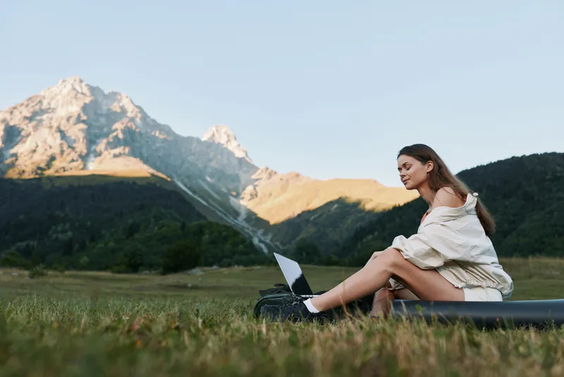 A woman sits on a grassy field with a laptop, smiling softly as she works outdoors, mountains rise behind her, creating a calm, productive, and inspiring nature office scene.