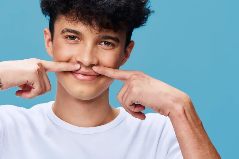young man making a mustache gesture with his fingers on blue background, playful expression, casual white t-shirt, curly hair, studio portrait
