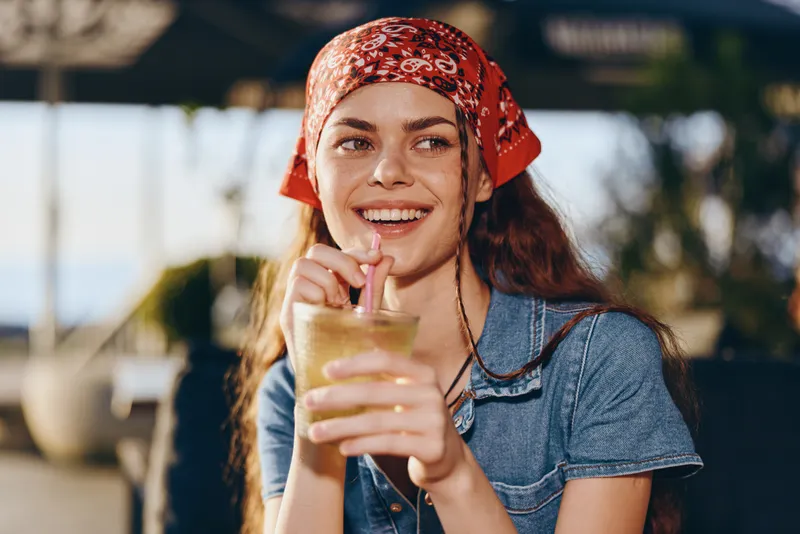 smiling woman drinking iced beverage outdoors wearing red headscarf and casual blue shirt under bright sunlight, enjoying refreshing moment with happy expression