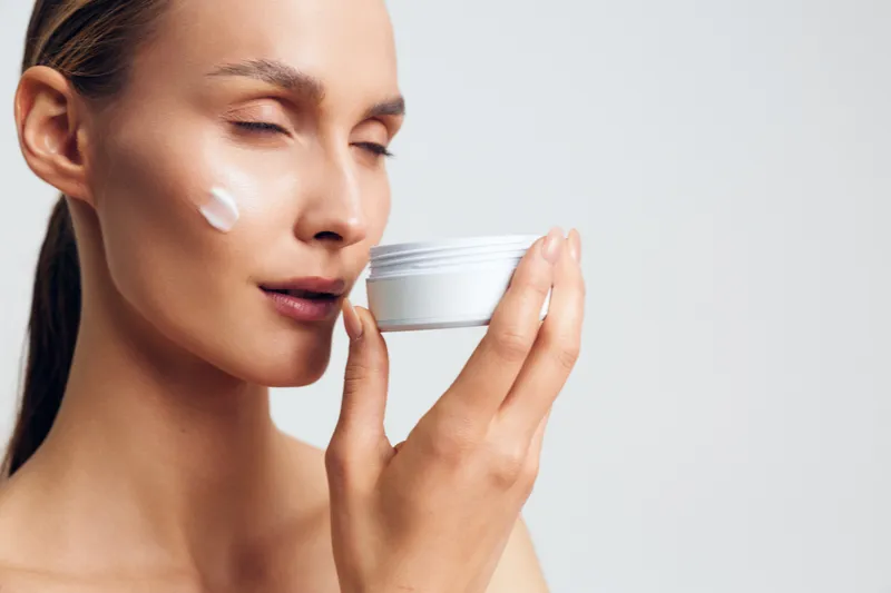 Young woman with clear skin applies moisturizing cream on her face while holding a white jar, enjoying a calm skincare routine against a plain light background.