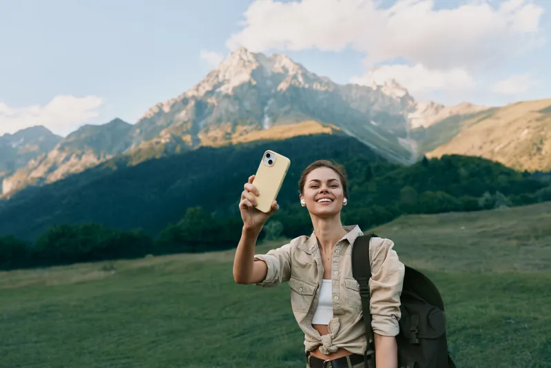 A young woman takes a selfie in a grassy valley with towering mountains behind her, capturing a spontaneous moment of adventure, confidence and freedom.
