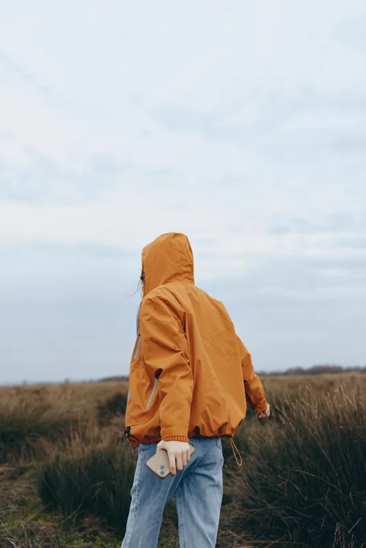Woman in orange hooded raincoat walking in a grassy field, casual jeans and phone in hand, cloudy sky behind, lifestyle outdoors, calm nature mood, soft natural light, fresh air, alone