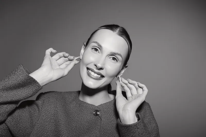 Smiling Caucasian woman with fair skin adjusts earrings wearing textured sweater. Studio portrait on grey background with soft lighting. Concept of fashion, beauty, and elegance.