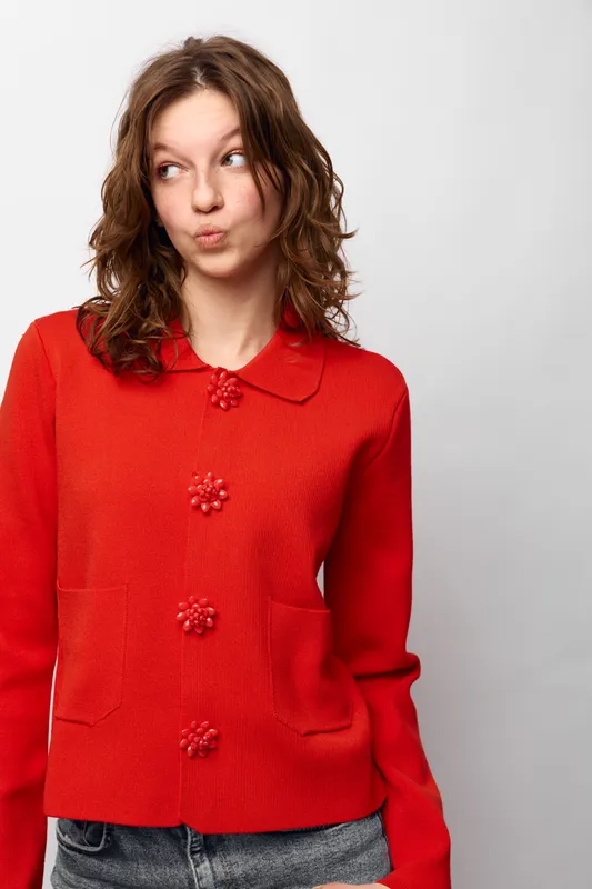 Young woman in red sweater with decorative buttons looks sideways with playful expression. Casual outfit and natural hairstyle on white background. Concept of fashion, youth, and personality.