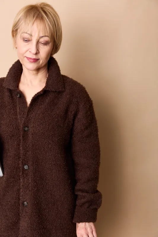 Elderly woman portrait indoors, wearing brown coat with soft fabric texture, standing against plain beige wall, serious face, looking down, vertical composition