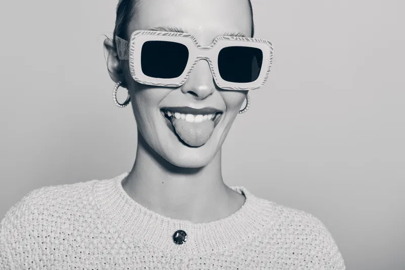 Smiling Caucasian woman with light skin wearing large sunglasses and knitted sweater shows tongue, expressing playful and confident mood in studio.