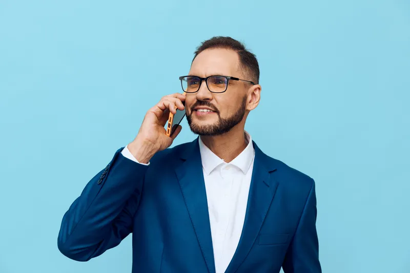 A professional man in a business suit stands against a solid blue background, exuding confidence and focus while on a mobile call.