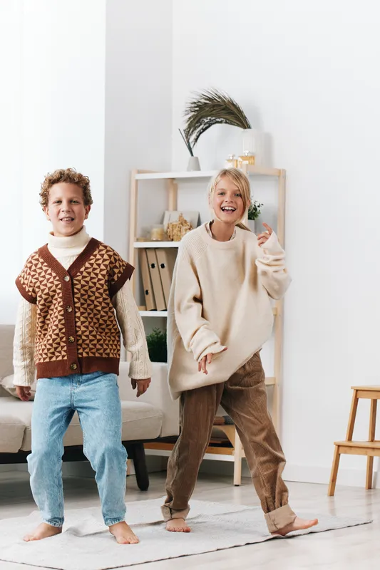 Two children dance playfully in a bright living room, wearing cozy knit sweaters and jeans, capturing joyful movement, warmth, and carefree afternoon energy.