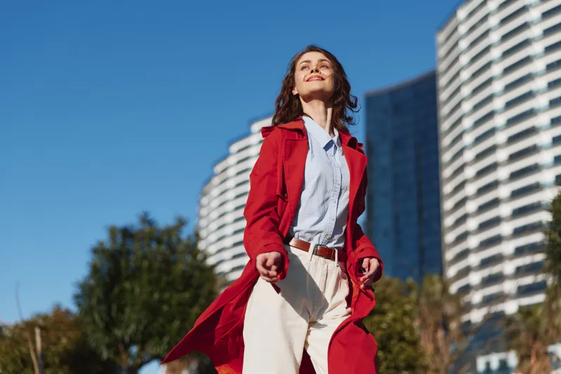 Vibrant woman in red coat stands outdoors against modern buildings, smiling confidently, wearing blue shirt and white pants, showcasing energetic urban street style.