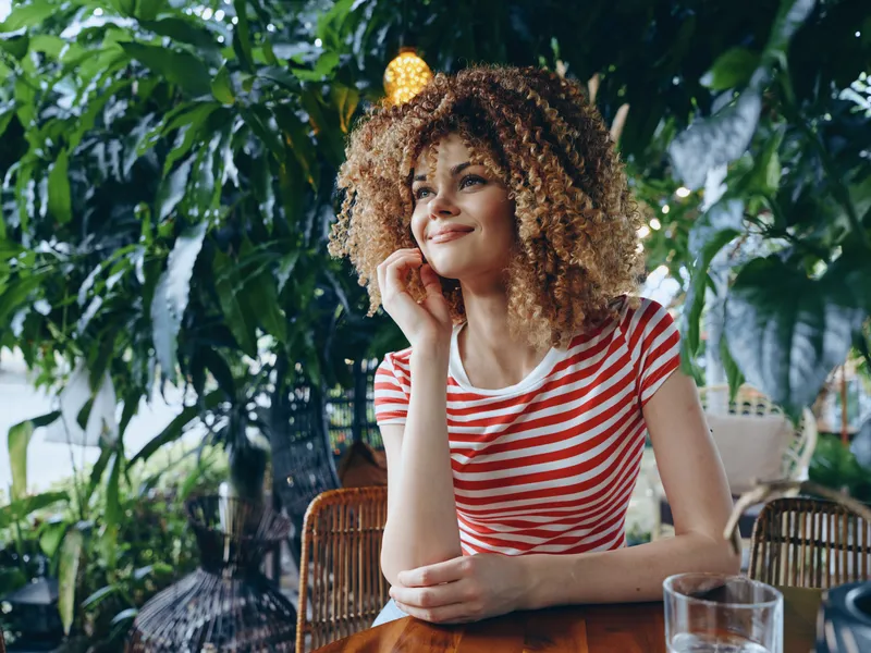 A thoughtful woman with curly hair sits at a wooden table in a lush cafe, wearing a red and white striped tee, soft natural light, calm candid moment.