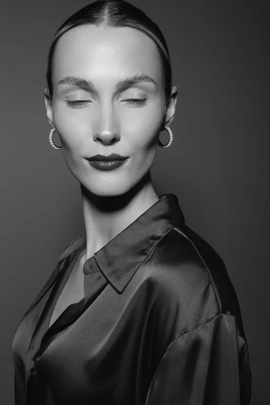 Portrait of a white woman with pale skin and closed eyes wearing hoop earrings and a satin blouse. Studio lighting creates a calm, elegant mood in black and white photography.