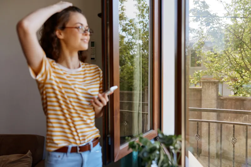 A woman in striped shirt stands by an open window, basking in daylight, lost in thought. A cozy home scene for lifestyle, relaxation, and simple living.