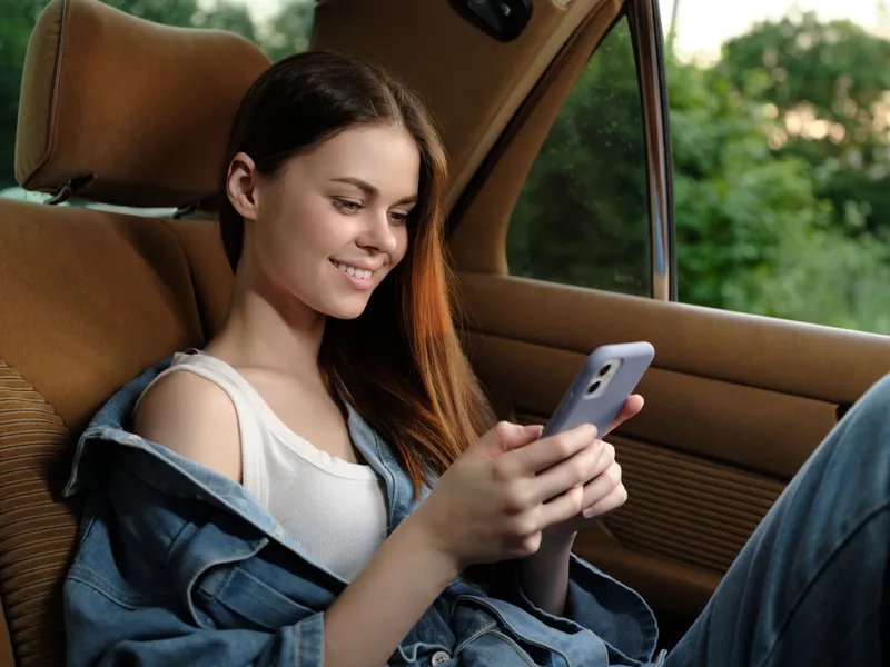 Young woman in casual denim attire smiling while using her smartphone in a car, reflecting a relaxed lifestyle and connection to technology.