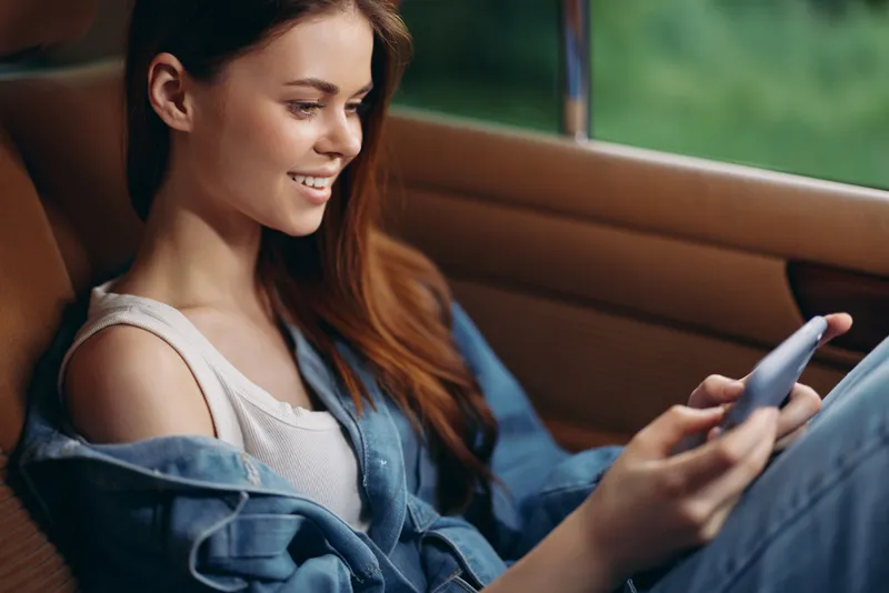 Smiling girl with long hair, sitting in a car, using a smartphone, wearing a denim jacket, enjoying the moment in a green outdoor background.