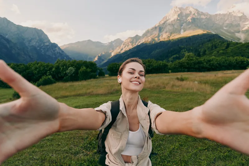 Young woman with a backpack takes a joyful selfie in an alpine meadow, mountains looming in the background, expressing freedom and happiness while casual travel attire invites viewers to join the