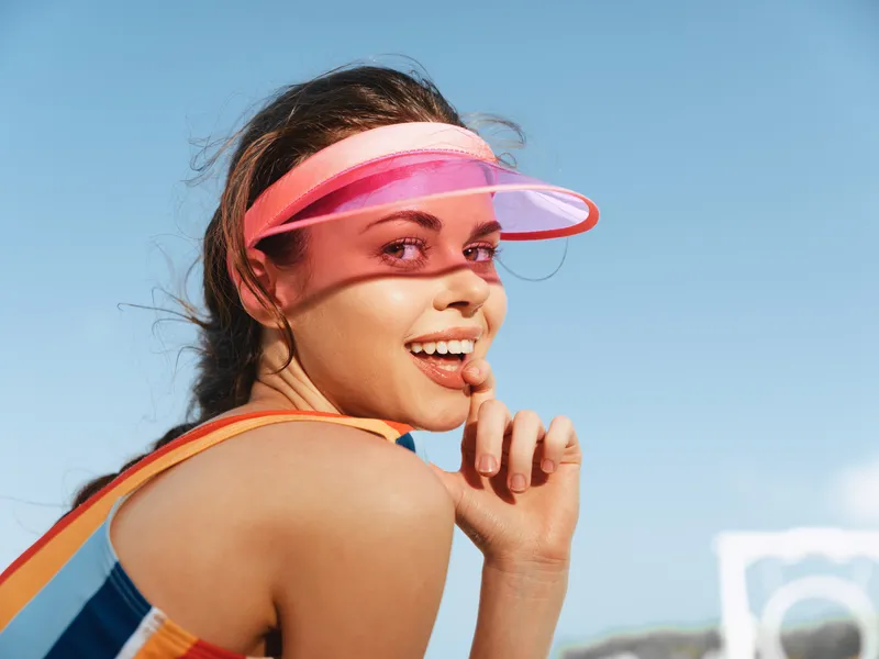 Young woman at the beach wearing a pink visor, laughing with a playful pose, sunlit skin, colorful sporty outfit, clear blue sky, a bright moment of carefree summer joy.
