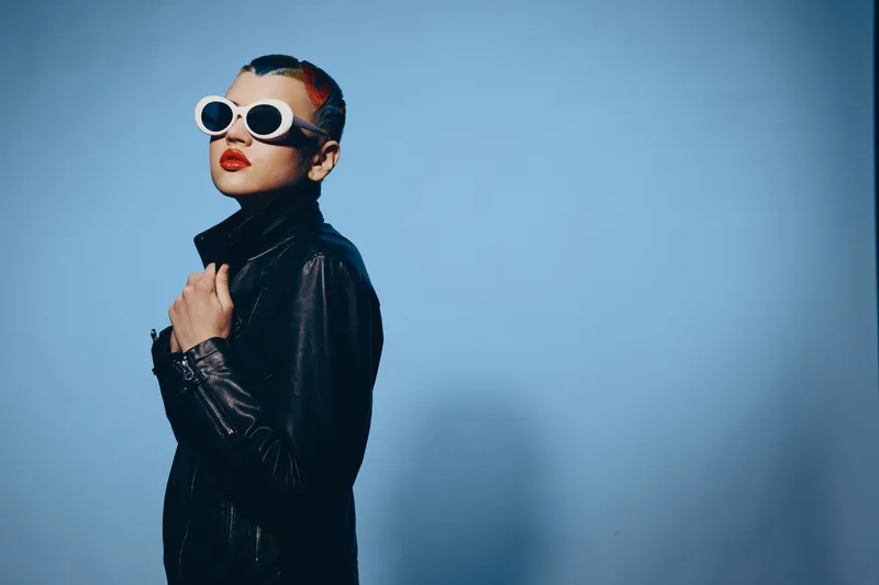 woman with sunglasses wearing black leather jacket stands confidently against blue background, featuring modern style and bold red lipstick in studio lighting.