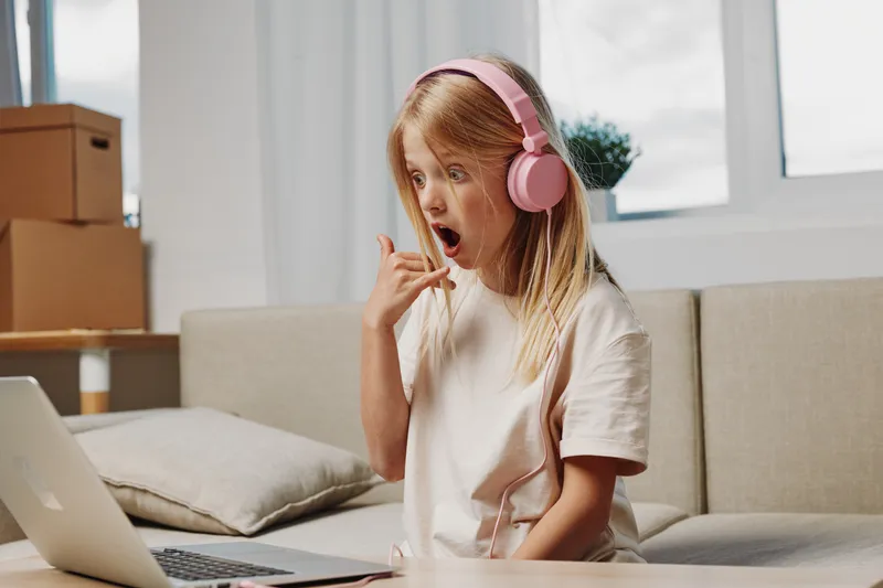 Surprised girl with pink headphones reacts to laptop screen at home, sitting on sofa in casual clothes. Concept of remote learning, communication, and technology use.
