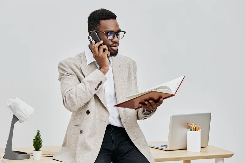 Professional young man with dark skin, wearing glasses and a beige blazer, standing in office, talking on mobile phone, holding open notebook, workspace with laptop, desk accessories, modern interior,