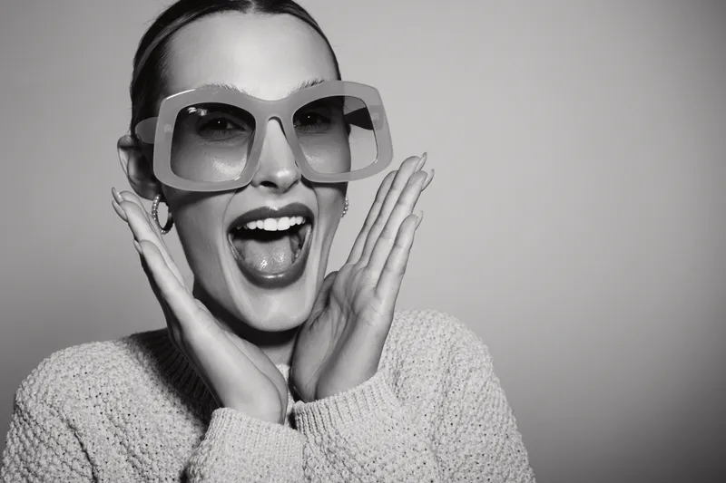Happy young Caucasian woman with fair skin wearing oversized sunglasses and sweater shows excited expression on grey background.
