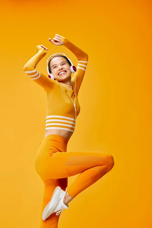 Woman in orange athletic outfit jumping midair against a bright orange background, wearing headphones, showcasing dynamic fitness and sport lifestyle with energetic movement.