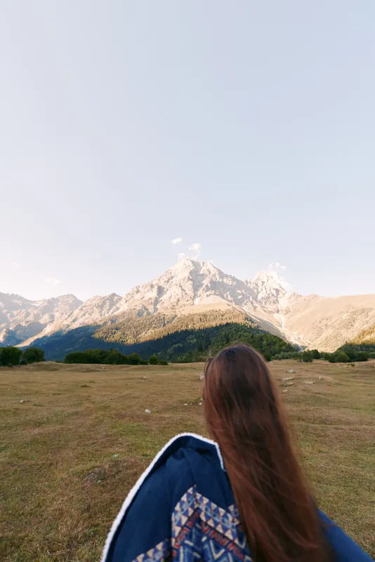 Mountain landscape with woman in meadow, outdoor hiking scene showing long hair and jacket from back, scenic peak and wide sky, travel and adventure mood in natural wilderness.