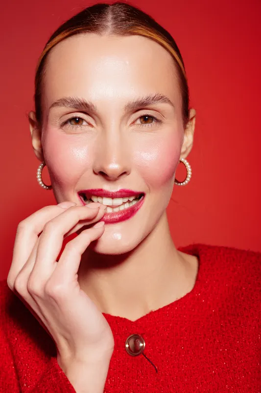 Close-up of smiling woman with red lipstick and pearl earrings wearing red sweater on red background. Concept of beauty, fashion, and confident female portrait.