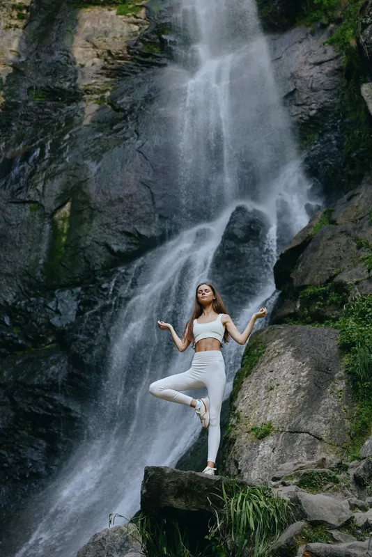 Woman practicing yoga in white clothes stands balanced on one leg near a cascading waterfall surrounded by rocks and green foliage, showcasing nature and wellness harmony.