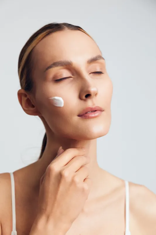 Woman closeup applies skincare cream on face, eyes closed, white product on cheek, smooth skin, beauty spa care, calm mood on neutral background.