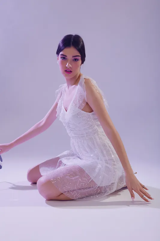 Young woman in a white lace dress kneels on the floor in a studio with soft purple lighting. Concept of beauty, fashion, elegance, and modern style for editorial or advertising.