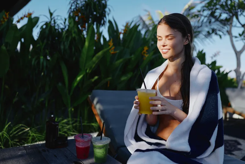 Casual lifestyle of a woman relaxing outdoors, wrapped in a towel, holding a glass of juice, sunny garden backdrop, bright colors, refreshing moment, enjoying a quiet morning breeze
