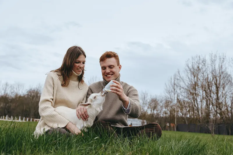 Happy couple outdoors with a playful goat kid in a grassy park, the man feeding the young animal while the woman strokes its fur, joyful lifestyle moment in a nature filled scene
