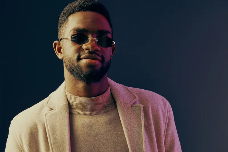 Stylish young man in sunglasses and a beige blazer, exuding confidence against a dark blue backdrop, perfect for fashion and lifestyle imagery
