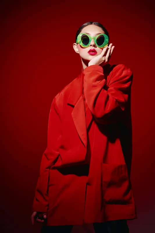 Fashion model with vibrant green sunglasses and bold red lips poses confidently in oversized red jacket on bright red background. Concept for beauty, creative fashion, and style.
