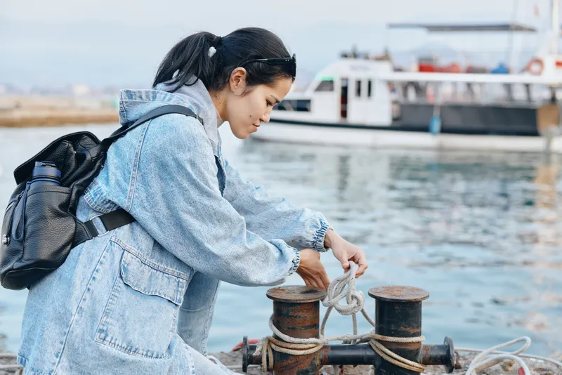 Harbor scene shows a woman in a denim robe tying rope to a bollard beside moored boats, preparing for docking and sailing operations on the quay during daylight