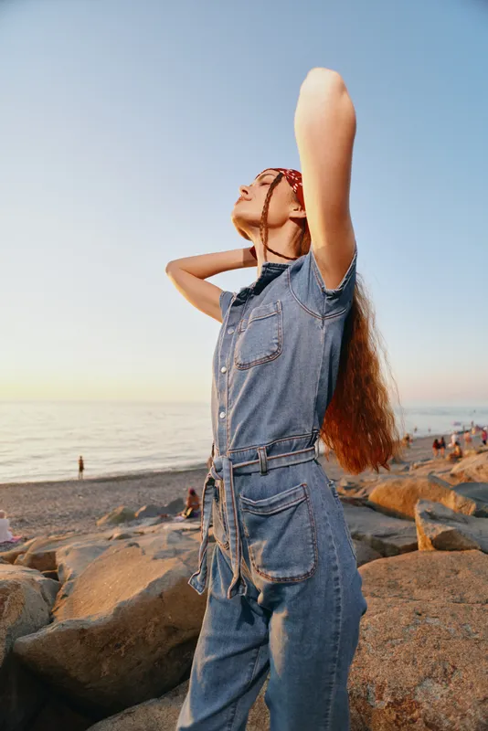 Lifestyle woman in modern boho-western denim street style enjoys sunny beach with film color tones and relaxed natural pose, capturing warm summer vibes.