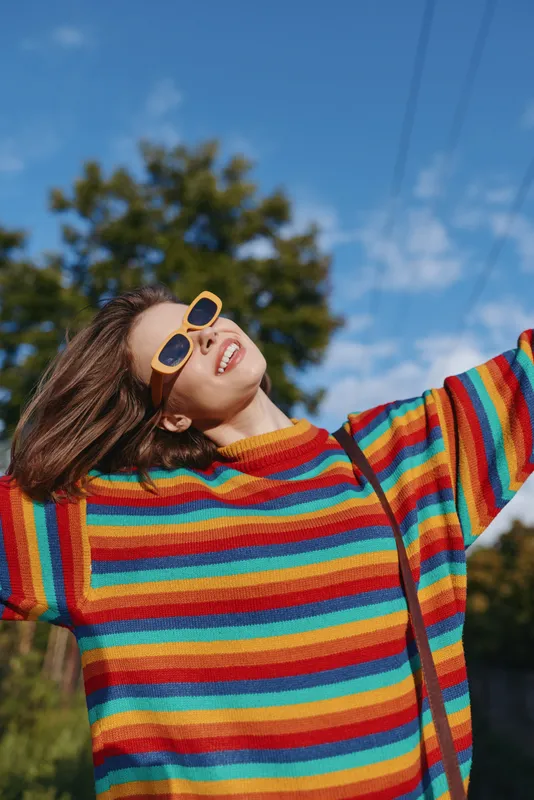 Woman in a rainbow sweater wearing yellow sunglasses, smiling and raising arms outdoors. Young adult casual outfit, joyful expression, leisure lifestyle portrait in bright sunny park.