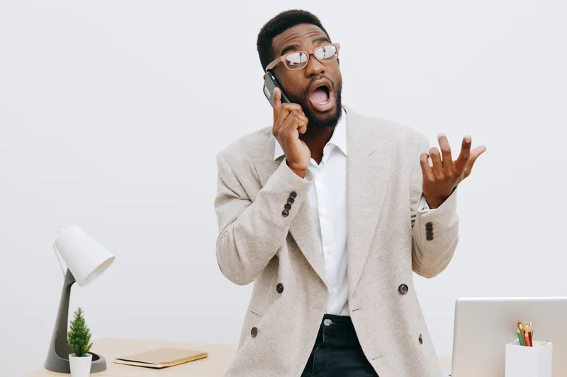 Professional young African American man wearing beige blazer, talking on mobile phone with surprised and expressive facial expression, standing at modern office desk with white background, concept of 