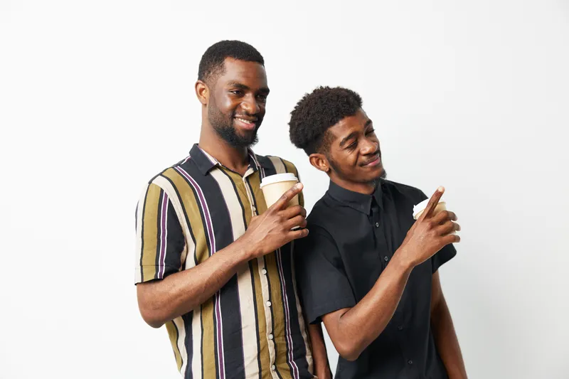 Two black men enjoying coffee together, sharing smiles and laughter, dressed casually in stylish shirts. Their camaraderie radiates joy and connection.