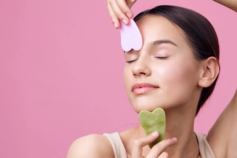 Young woman using facial massage tools for skincare on a pink background, promoting beauty and self care with positive emotions