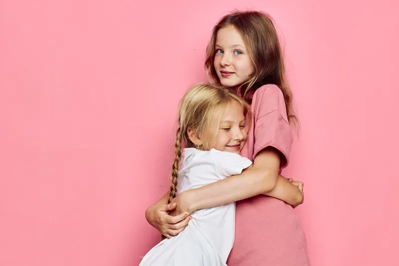 Two happy girls hugging against a pink background. Older girl looks at camera while younger girl smiles with eyes closed.