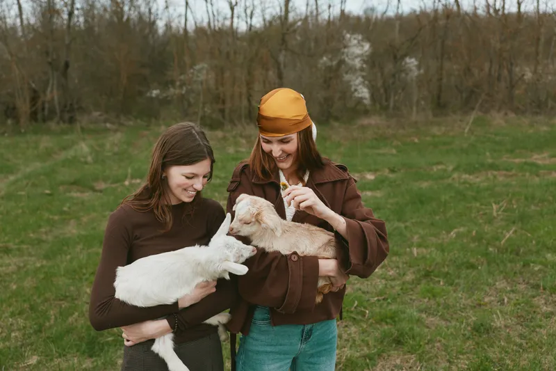 Two young women enjoy a casual outdoor moment holding a baby goat and a kid while smiling and wearing cozy autumn clothes in a natural grassy field.