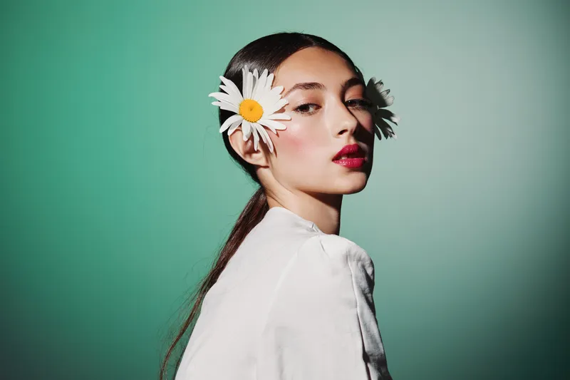 Portrait of a young woman with smooth skin posing with daisy flowers in her hair against green background. Concept of beauty, creative fashion, fresh, elegant.