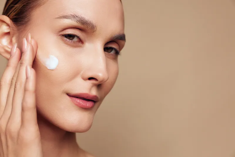 Young woman applies facial cream on her cheek in a close up portrait with beige background, calm expression, natural light, skincare routine in progress.